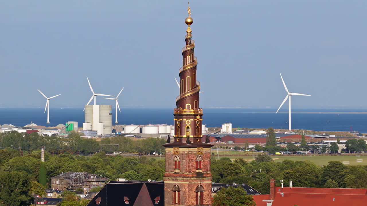 Aerial drone view of the Church of Our Saviour with its golden spiral tower, set against a backdrop of wind turbines and the sea in Copenhagen, Denmark