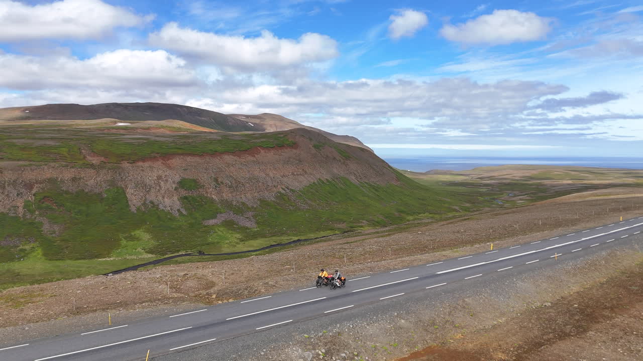 Vista aerea da drone di due cicloturisti che pedalano in salita su una strada asfaltata attraverso un paesaggio montuoso isolato a Langanesbyggð, in Islanda, sotto un cielo parzialmente nuvoloso