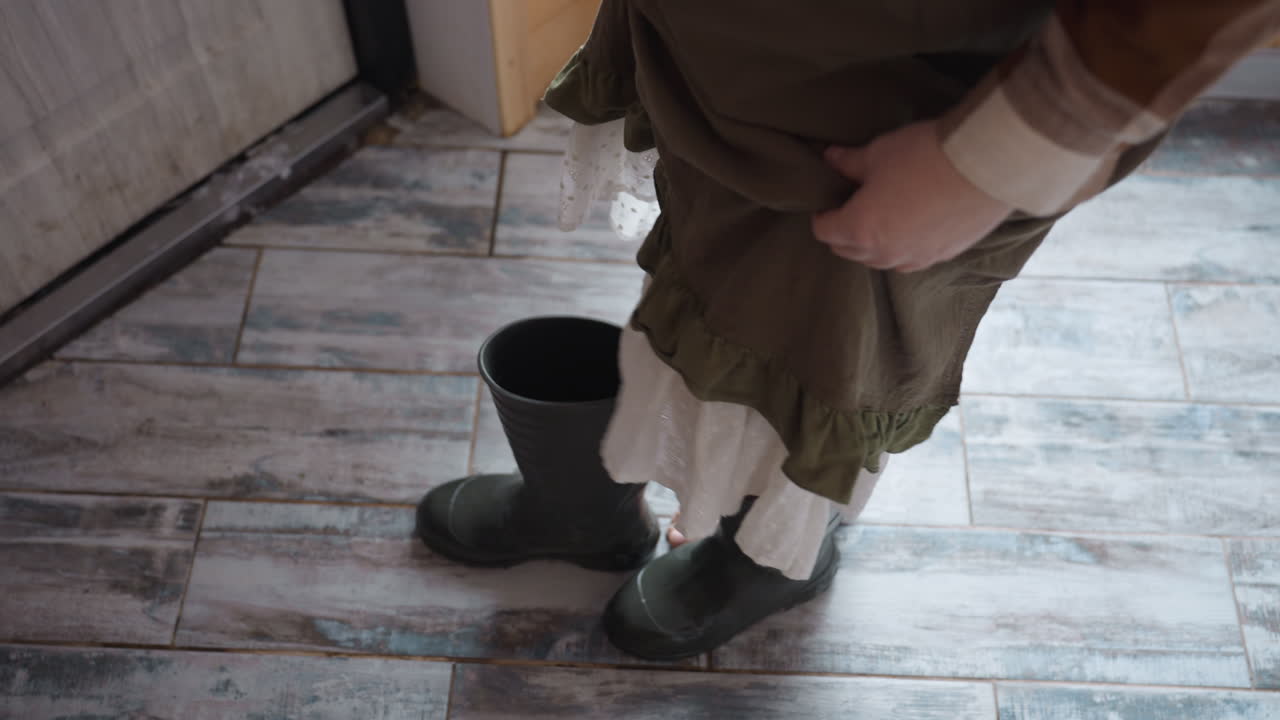 Green activist stepping barefoot into green snow boots on rustic tiled floor, dress hem ruffling, preparing for winter outdoor walk under soft indoor winter light casting gentle shadows