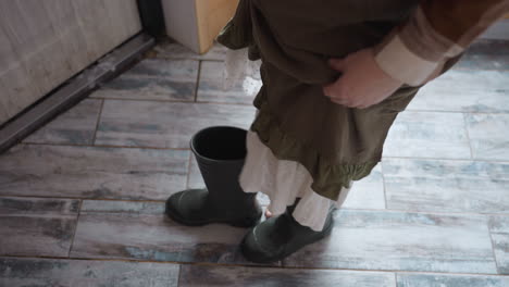 green activist stepping barefoot into green snow boots on rustic tiled floor, dress hem ruffling, preparing for winter outdoor walk under soft indoor winter light casting gentle shadows
