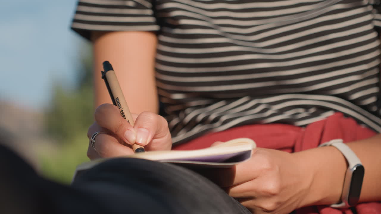 White poet journaling on lap outdoors, close framing of hands, pen and notebook, striped top and red jacket detail, rings and smartwatch visible, warm natural light and quiet introspective tone