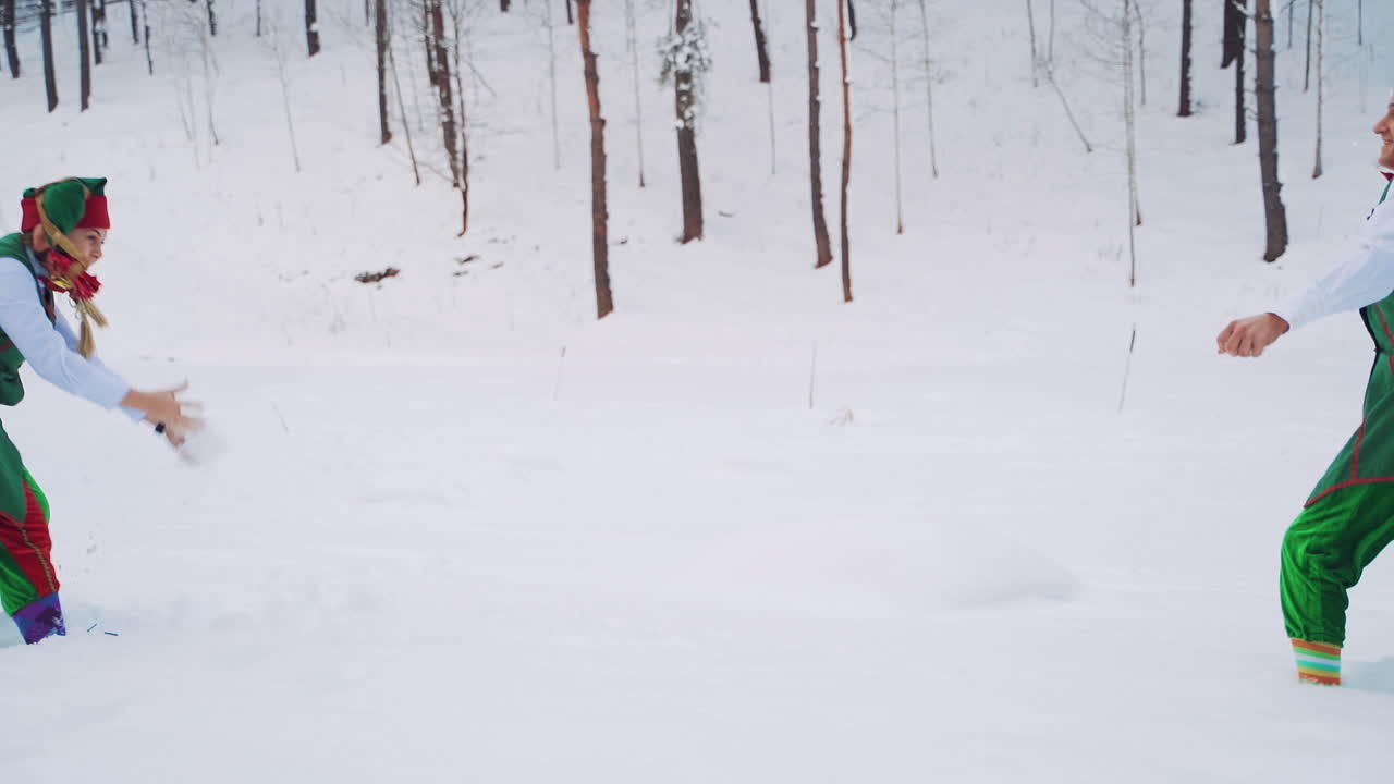 Young elf girl in green suit and a hat is running in the snow joyfully. Two friends elves are playing snowballs together in winter on the background of the forest. Slow motion.