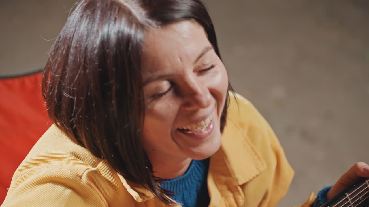 Donna in sedia da campeggio che suona l'ukulele, primi piani di espressioni gioiose e giacca calda, sedia da campeggio pieghevole rossa visibile, risate sincere e canto sommesso, accordatura e delicato fingerpicking, intimo