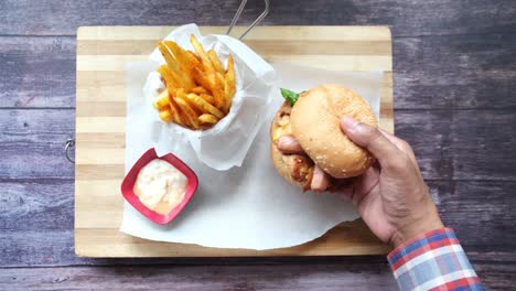 hand holding a burger with fries and a side of sauce on a wooden cutting board.