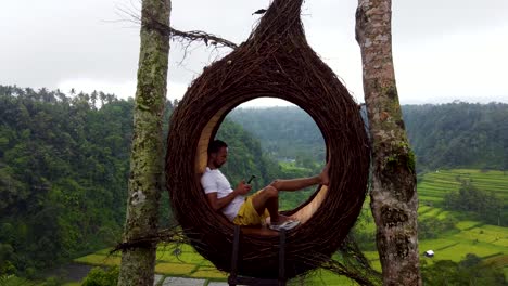 Aerial-view-:-A-young-tourist-sitting-inside-a-bird-nest-instagram-photo-spot-on-a-hill-with-a-stunning-view-to-green-rice-terraces-and-fields-operating-a-drone-to-take-a-selfie