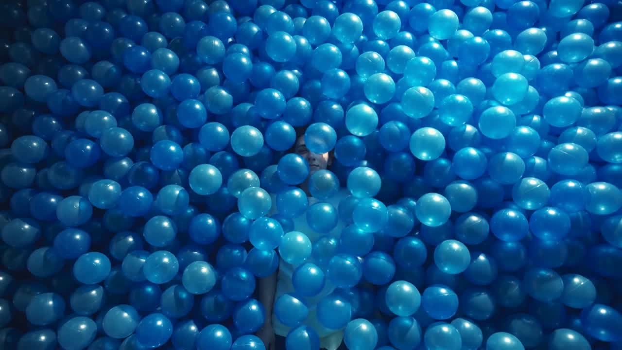Forskydende blå bolde afslører en voksen kvinde, der ligger tilbagelænet i et boldbassin, iført en lysfarvet top.
