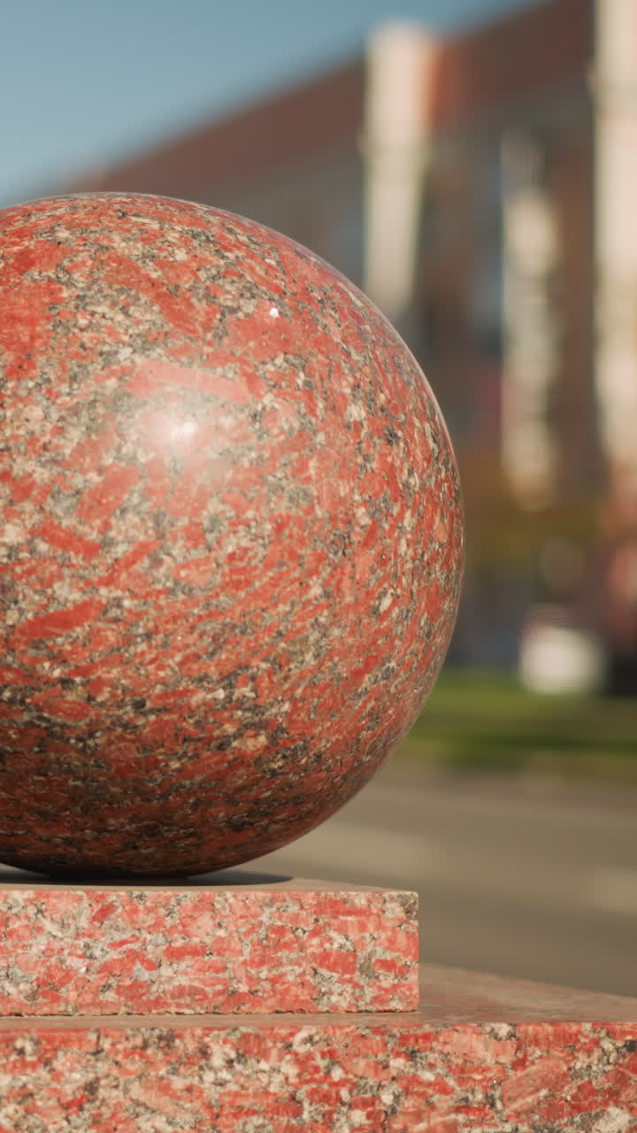 Red granite sphere on urban pedestal bathed in bright midday sunlight, shallow depth of field with blurred cars and modern building facade in background, detailed speckled texture and polished sheen