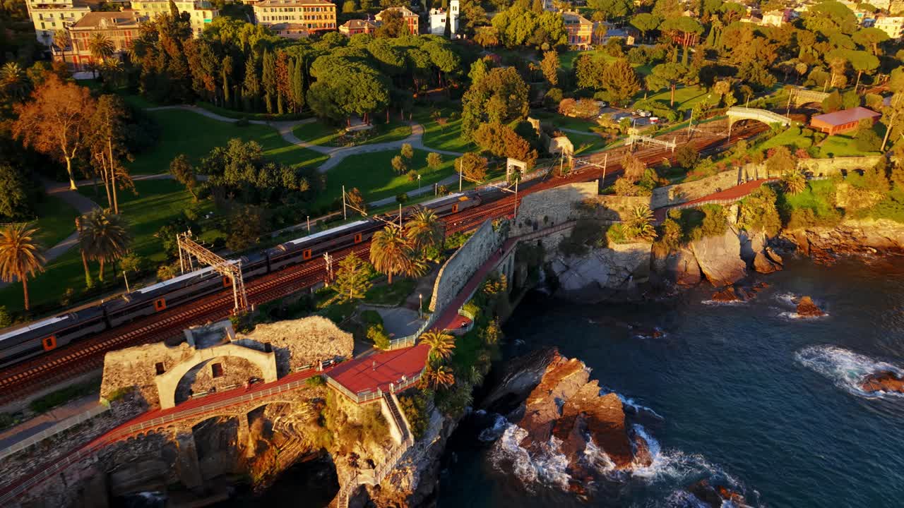 Golden hour drone shot of Genoa's coastline, showing lush parks, train tracks, and the sea