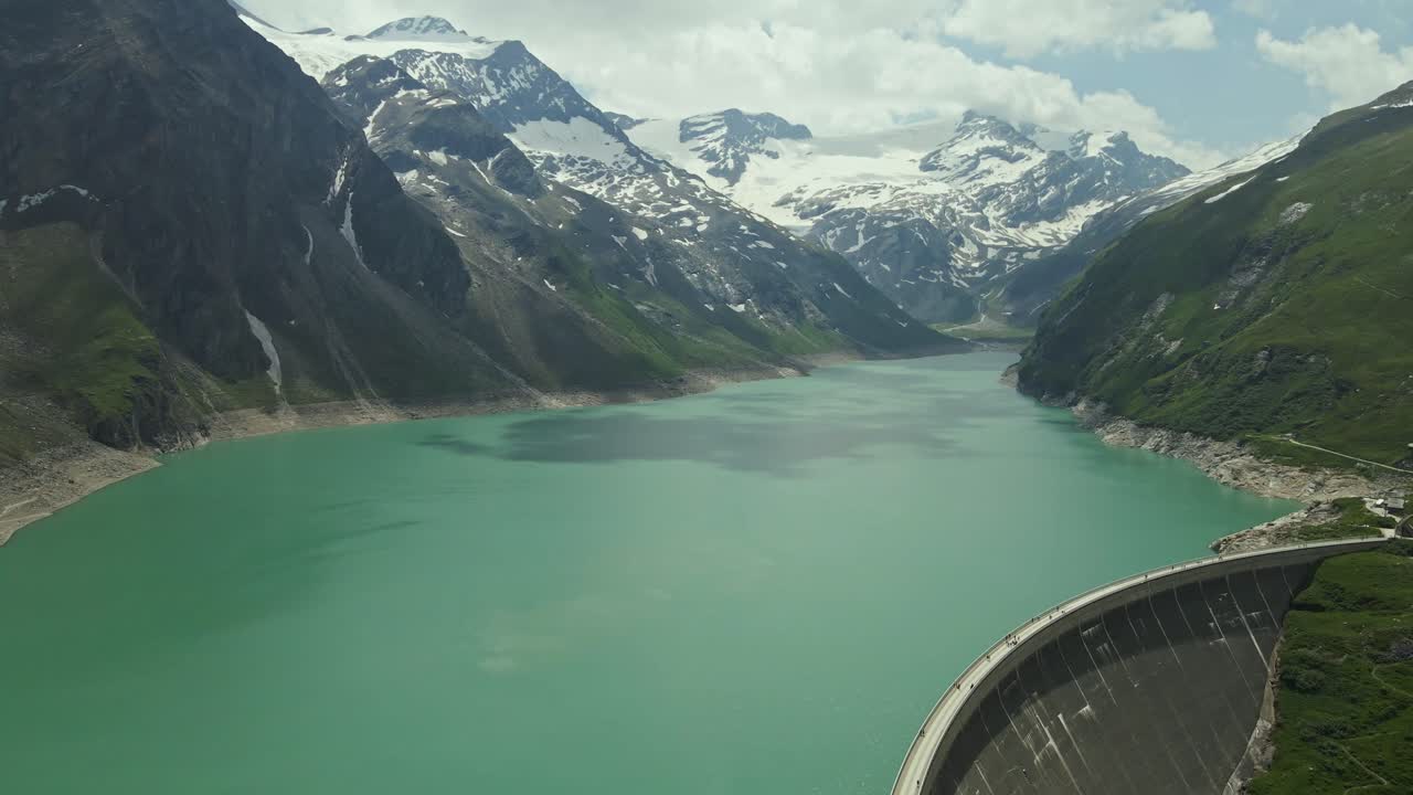 Aerial view of a turquoise reservoir framed by massive dam walls in a mountain valley. Snow-covered alpine peaks rise in the background, contrasting with the green slopes and the calm water surface.