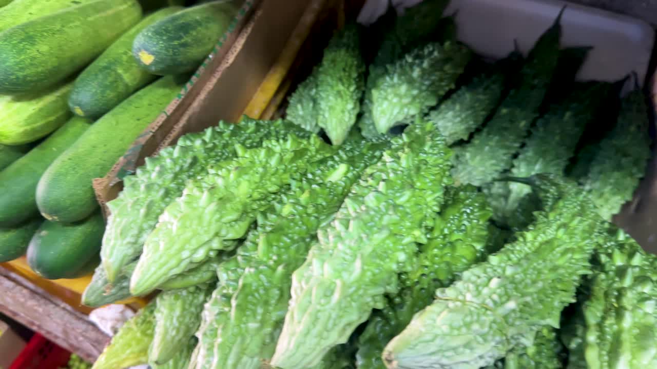 Close-up views of fresh bitter gourd and cucumbers arranged in crates at a vibrant vegetable market, captured with smooth camera movement and bright lighting