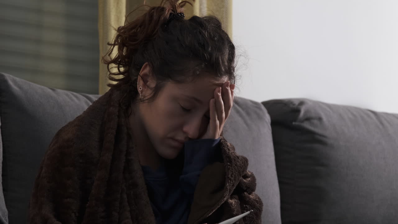 Giovane donna malata sul divano di casa, con un termometro. Pillole, fazzoletti e una tazza sul tavolo.