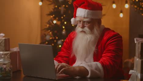 Weihnachtsmann-Benutzt-Laptop-Im-Wohnzimmer.-Porträt-Des-Nachdenklichen-Weihnachtsmanns,-Der-Mit-Modernem-Laptop-In-Dekoriertem-Haus-Tippt.-Hochwertiges-4K-Filmmaterial