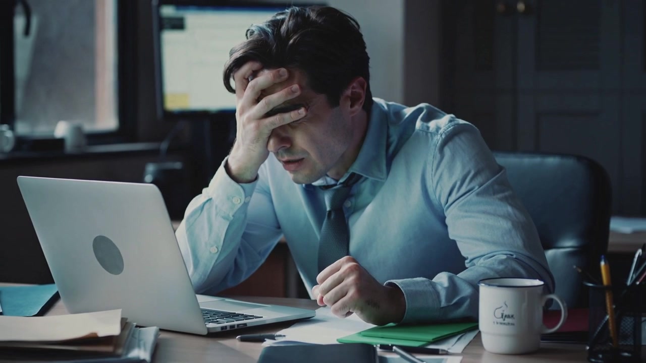 Un uomo chino in camicia blu e cravatta scura preme la fronte sulla scrivania dell'ufficio, con un laptop che segnala il carico di lavoro.