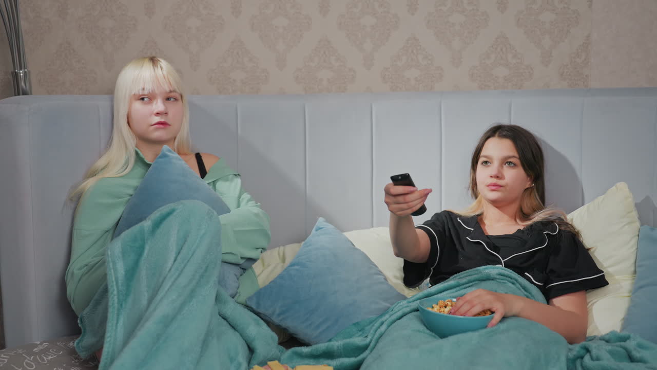 Two teenage girls in pajamas relaxing on bed watching movie, one girl holds remote control trying to change channel while eating popcorn, other hugs pillow with serious expression
