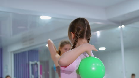 vista parcial del director del gimnasio instruyendo a las damas jóvenes en la flexibilidad del brazo con vista trasera de la dama luchando para alcanzar con la otra mano, en un gimnasio bien iluminado con espejos