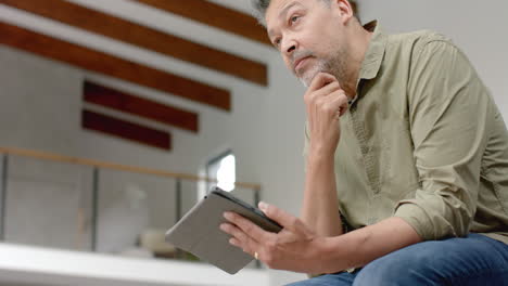 thoughtful senior biracial man sitting on table using tablet at home, copy space, slow motion