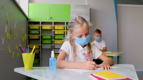 little girl with face mask in class and hand sanitizer on table doing exercises in a notebook. covid-19 pandemic. child back to school after coronavirus lockdown.