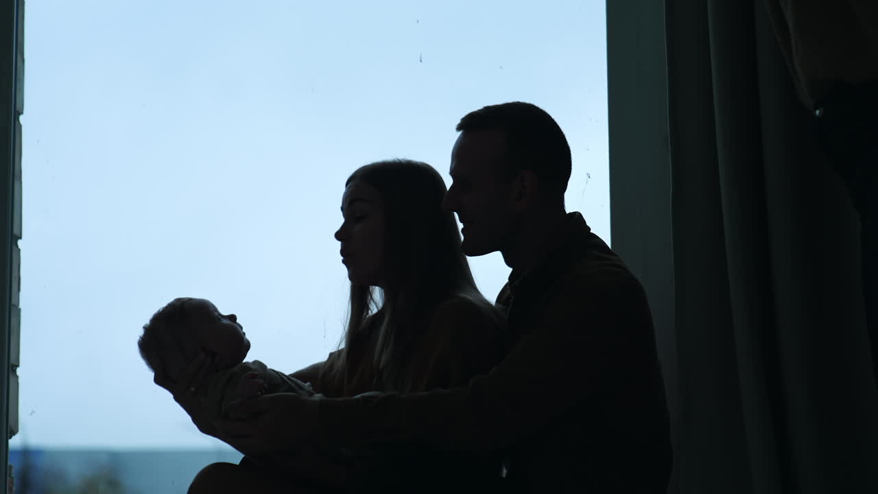 Man and woman sitting at the window-sill waving a baby. Parents enjoying their parenthood.