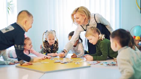 children learn at round table with smiling teacher in class. little kids taking cards to themselves from table during english lesson indoors.