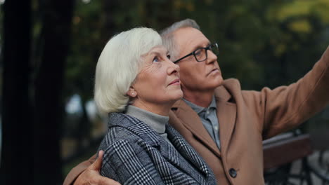 elderly couple sitting on the bench in the park, the man points upwards showing it to his wife