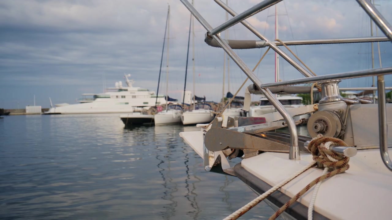 Close-up da proa de um veleiro, âncora, cordas e grades de metal na marina Port Vauban em Antibes, com iates e águas calmas do Mediterrâneo ao fundo.