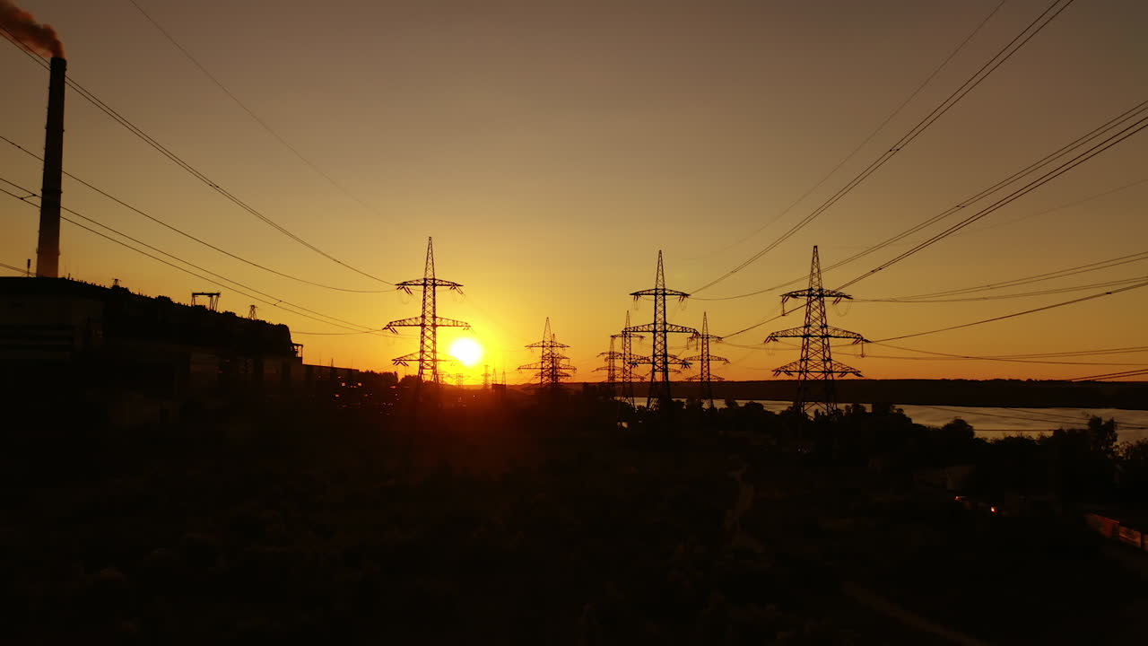 Silhouette of electric towers against orange sunset. High-voltage electric lines among the evening nature. Electricity efficiency. Transmission lines and factory.