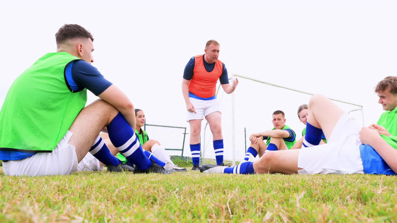Male and female soccer players sitting and listening to coach giving instructions on pitch