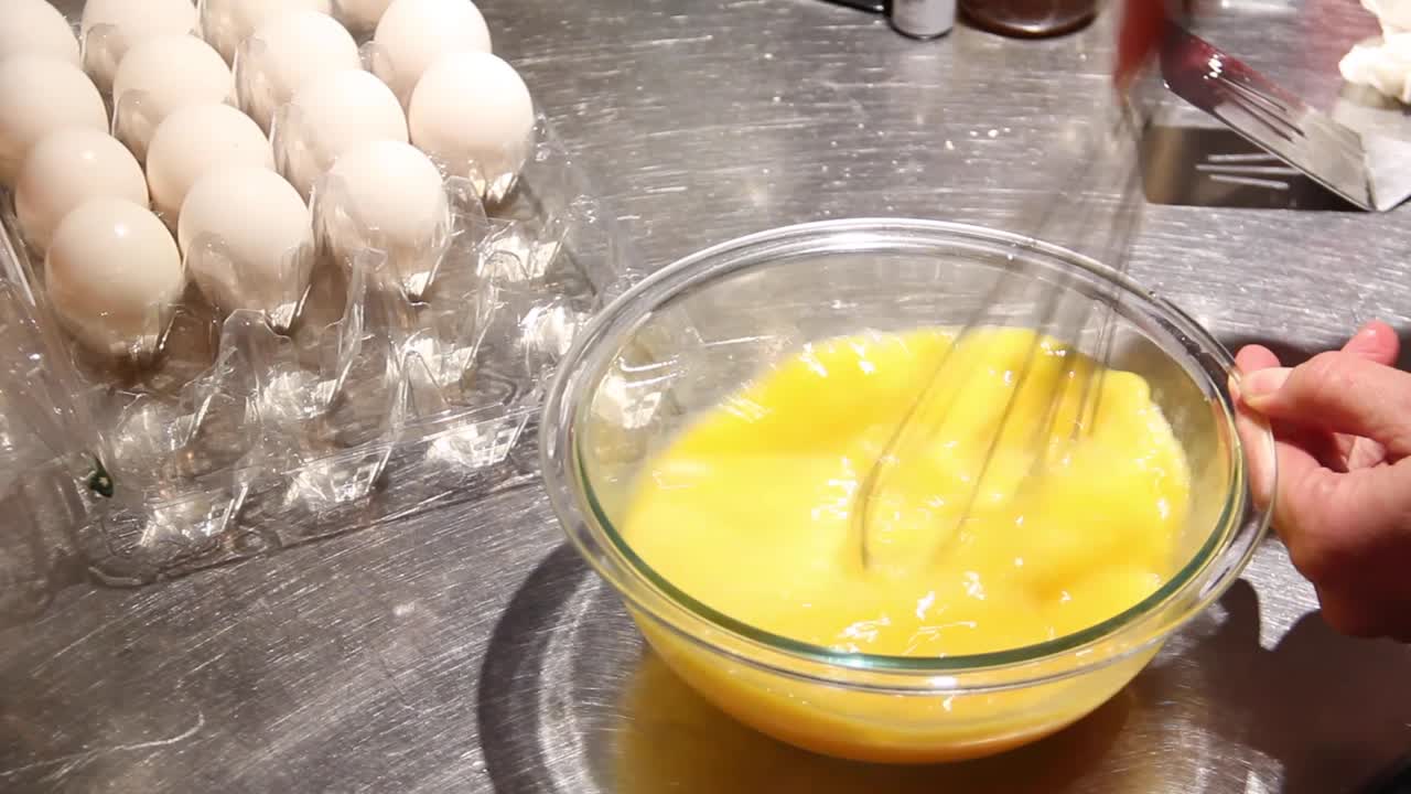 Eggs in a bowl on a clean stainless steel countertop are beaten with a whisk