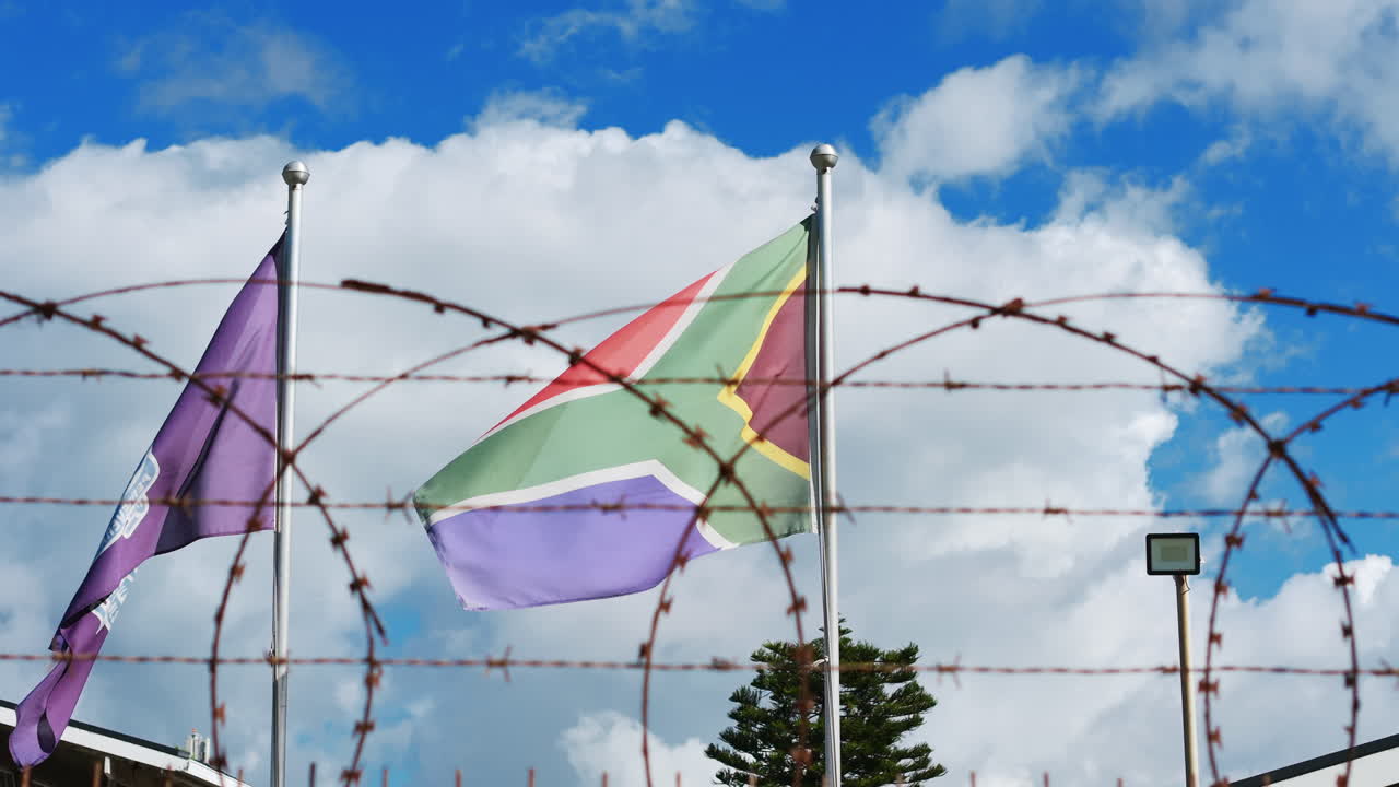 The South African flag flutters behind barbed wire under a vivid blue sky, capturing a powerful contrast of resilience, identity, and guarded reality in Cape Flats