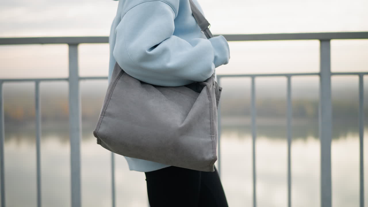 Partial view of young woman wearing blue sweater and black leggings, holding bag while walking calmly, background features iron railing and serene river view