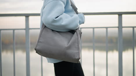partial view of young woman wearing blue sweater and black leggings, holding bag while walking calmly, background features iron railing and serene river view