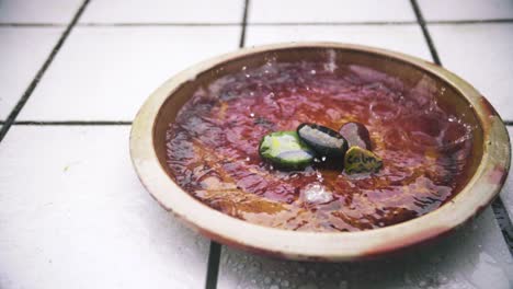 Close-Up-Pan-of-Slow-Motion-Rain-flooding-a-brown-flower-clay-jar-with-white-tiles-in-background-and-stones-written-the-words-calm-and-luck-on-them