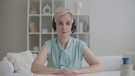 portrait of online consultant woman with headphones on head sitting at table and talking to camera