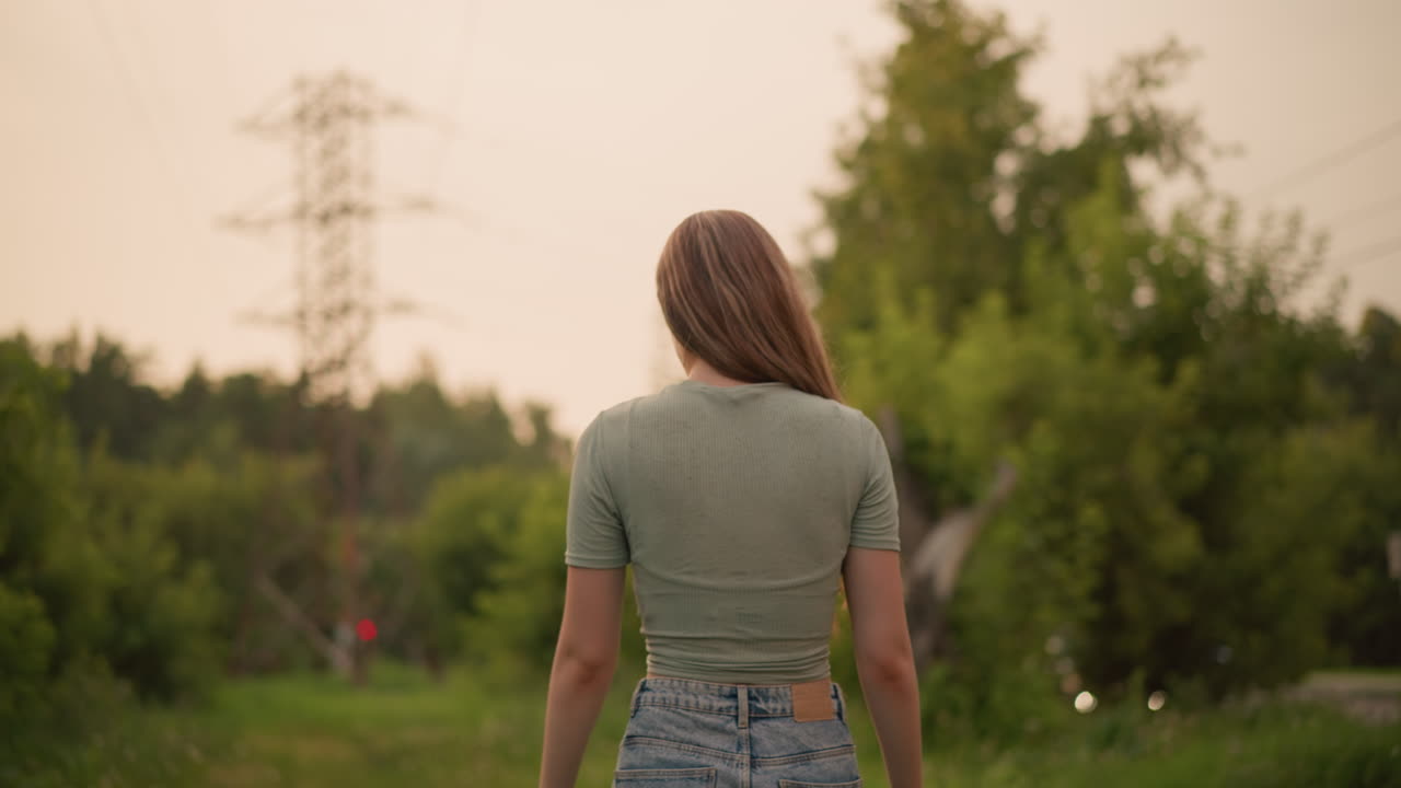 잔디로 된 길을 따라 걸어가는 젊은 여성, 그녀의 머리카락이 앞으로 흐르고, 전선 에서 조용하고 자연스러운 장면을 만들고, 먼 차와 초록색을 특징으로하는 흐릿한 배경