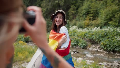 foto de una chica tomando una foto de su amiga que está de pie con una bandera lgbt en una camiseta blanca contra el telón de fondo de un bosque verde