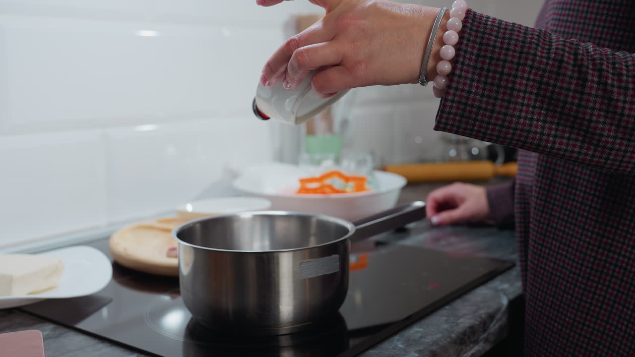 Close-up of hand in checkered sleeve adorned with beaded bracelet pouring honey into pot on electric stove, modern kitchen background with ingredients, marble countertop and cooking utensils