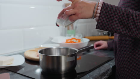 primer plano de la mano en la manga a cuadros adornada con una pulsera de cuentas vertiendo miel en la olla en la estufa eléctrica, fondo de la cocina moderna con ingredientes, encimera de mármol y utensilios de cocina