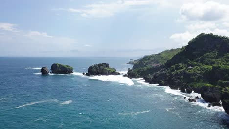 drone shot of cliff with rocky island on the beach,hit by waves during sunny day - timang island, yogyakarta, indonesia