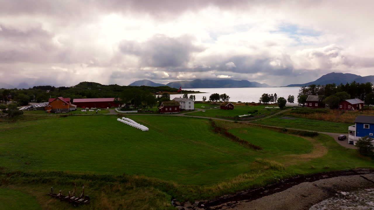 Aerial drone view of Kvitnes Gård, a historic farm and restaurant in Vesterålen, Norway. Traditional red and white buildings with sod roofs sit amidst green fields near the fjord under a cloudy sky