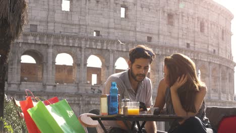 happy young couple tourists using smartphone sitting at bar restaurant in front of colosseum in rome at sunset with coffee shopping bags smiling having fun texting browsing and sharing pictures