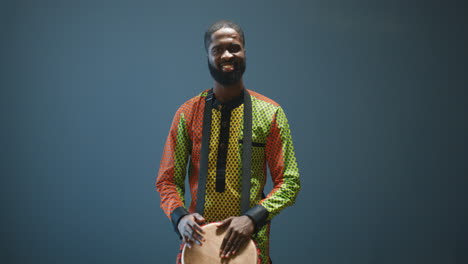 portrait of young cheerful african american man in traditional clothes playing a drummer and smiling at camera