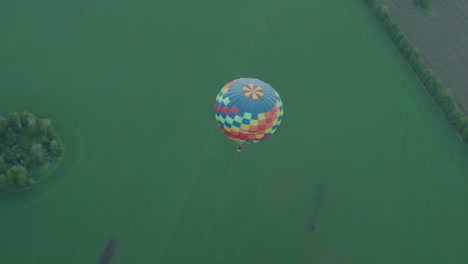 colorful checkered hot air balloon carrying two passengers drifts silently over vast green farmland at sunset, aerial perspective reveals winding river and tree lines