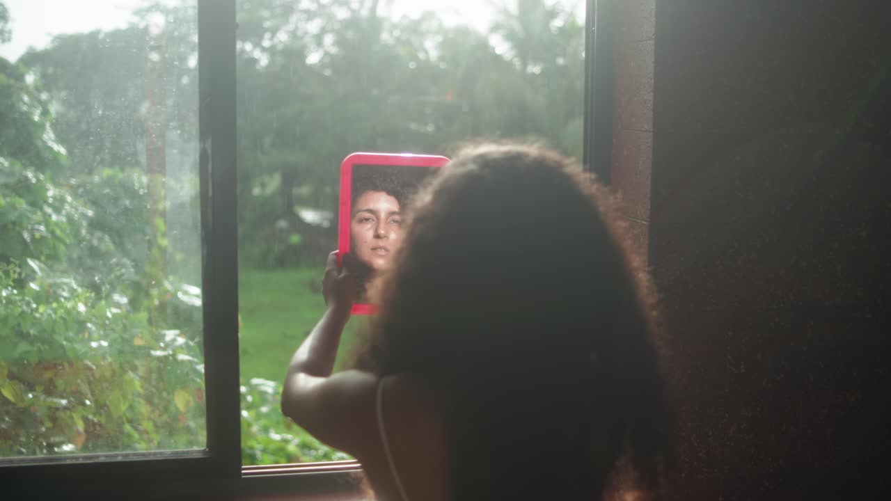 Fixed shot of woman from behind holding a red mirror at a window, reflecting her face with garden greenery outside