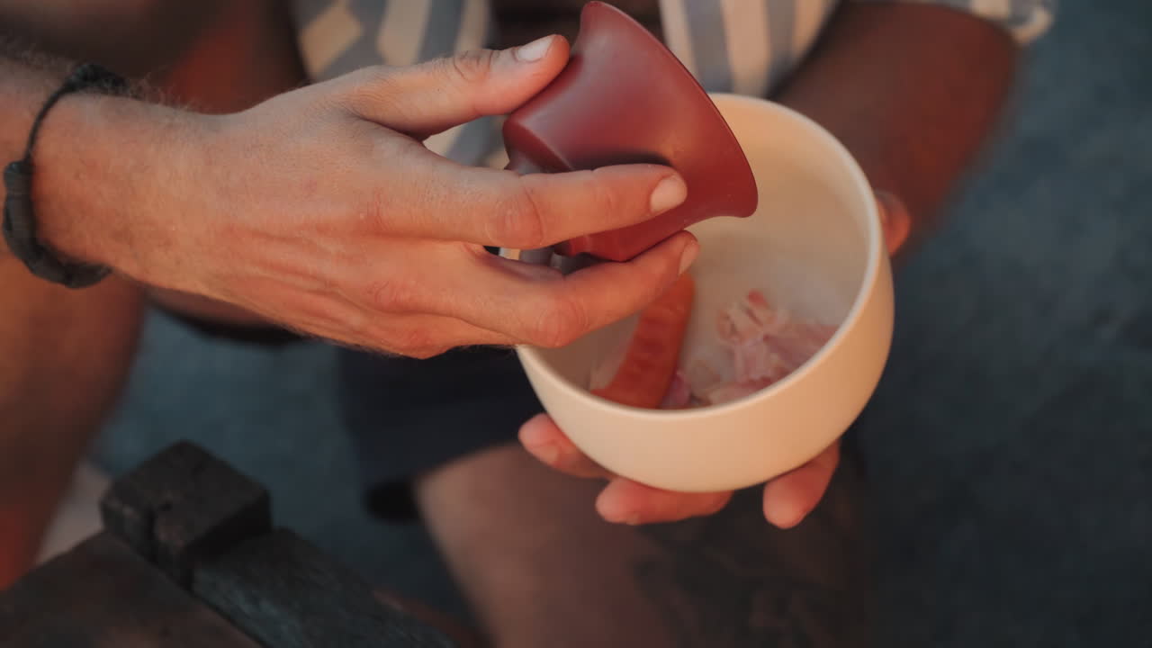 Persona che versa il tè su un vassoio da spiaggia al tramonto, tazze di argilla e foglie sfuse su un vassoio nero, un lento rituale consapevole condiviso da una coppia, caldo bagliore dorato, gambe nude, primo piano di mani che eseguono una cerimonia delicata