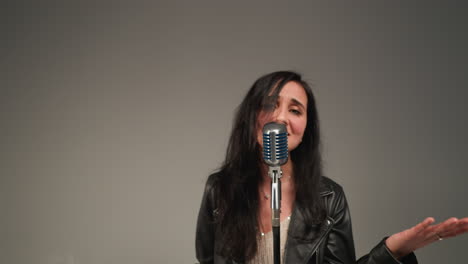 a vocalist in a black leather jacket sings with expressive hands and eyes open as the camera zooms in and out. in front of a vintage microphone against a grey background