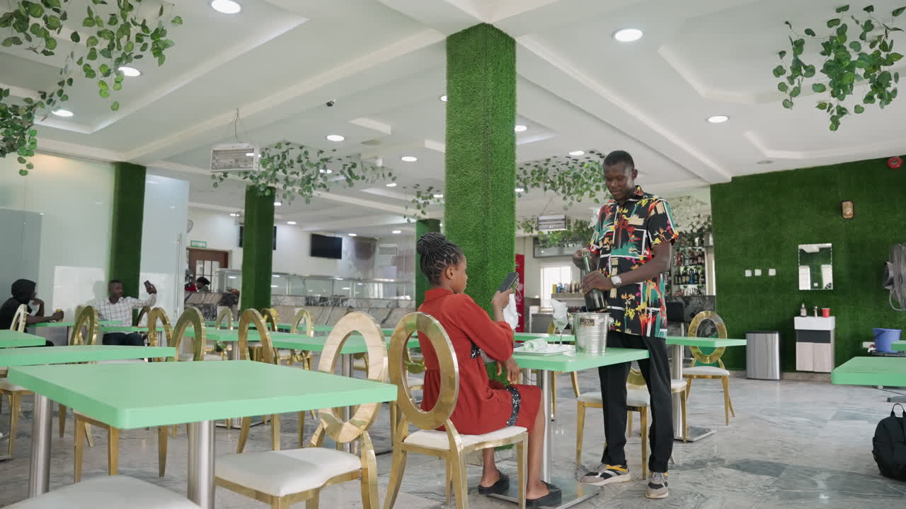 Coppia afroamericana a un appuntamento romantico al caffè, la donna seduta in abito rosso sorride mentre l'uomo in piedi offre un piccolo regalo e alza il bicchiere, la camicia a fantasia contrasta con l'interno lussureggiante coperto di piante, moderno e luminoso