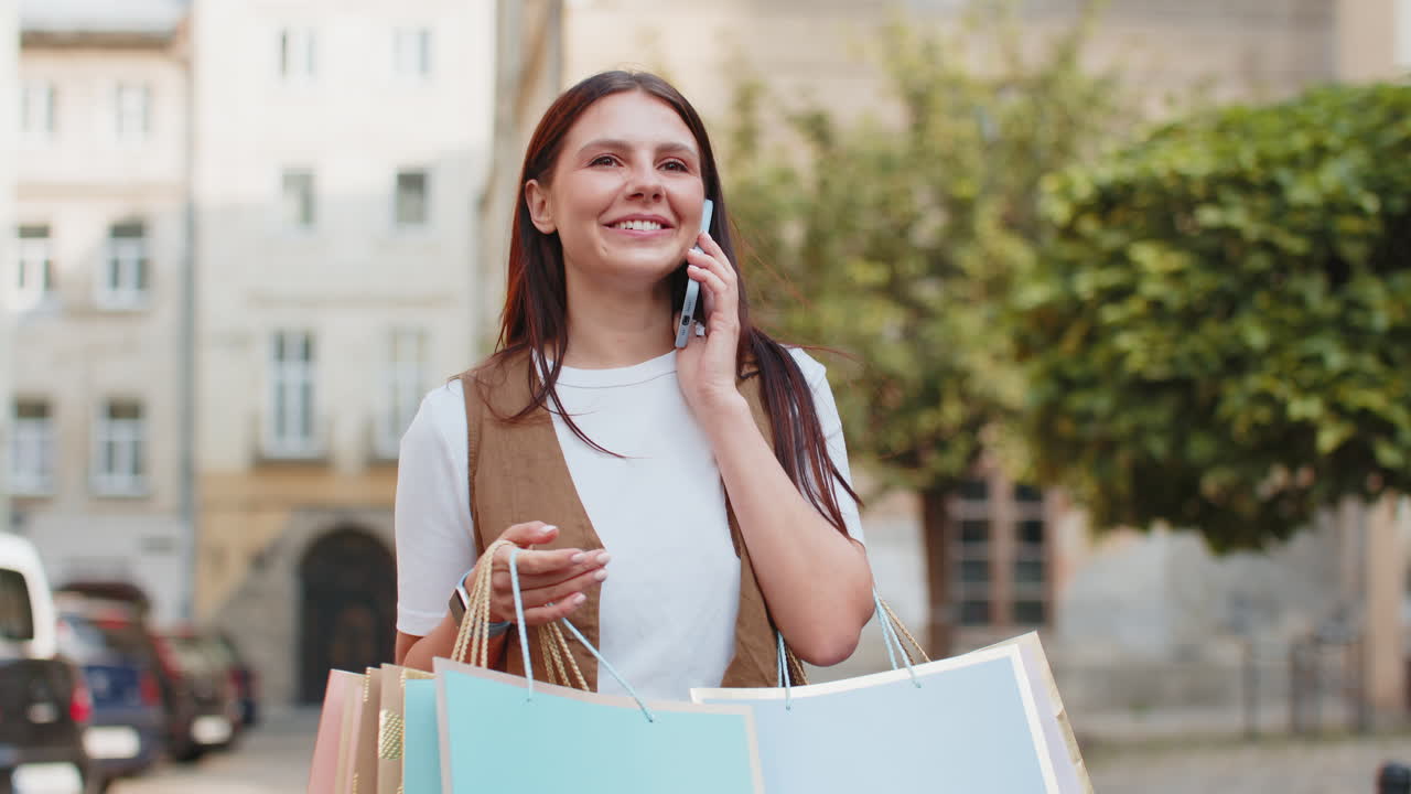 joven feliz adicta a las compras que lleva bolsas de compras mientras habla a través de un teléfono inteligente en la calle de la ciudad