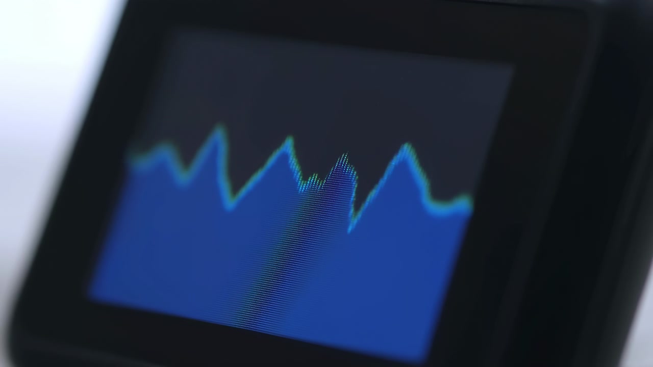 Angled close-up of an electronic display showing a blue fluctuating line chart against a dark background, with soft light and shallow focus highlighting the