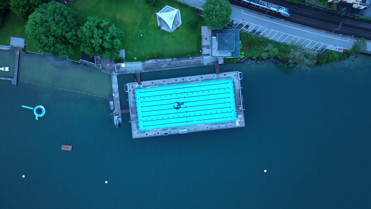 Deserted swimming pool along shores of lake or river, train passing, sunset light, blue hour. Aerial top-down ascending