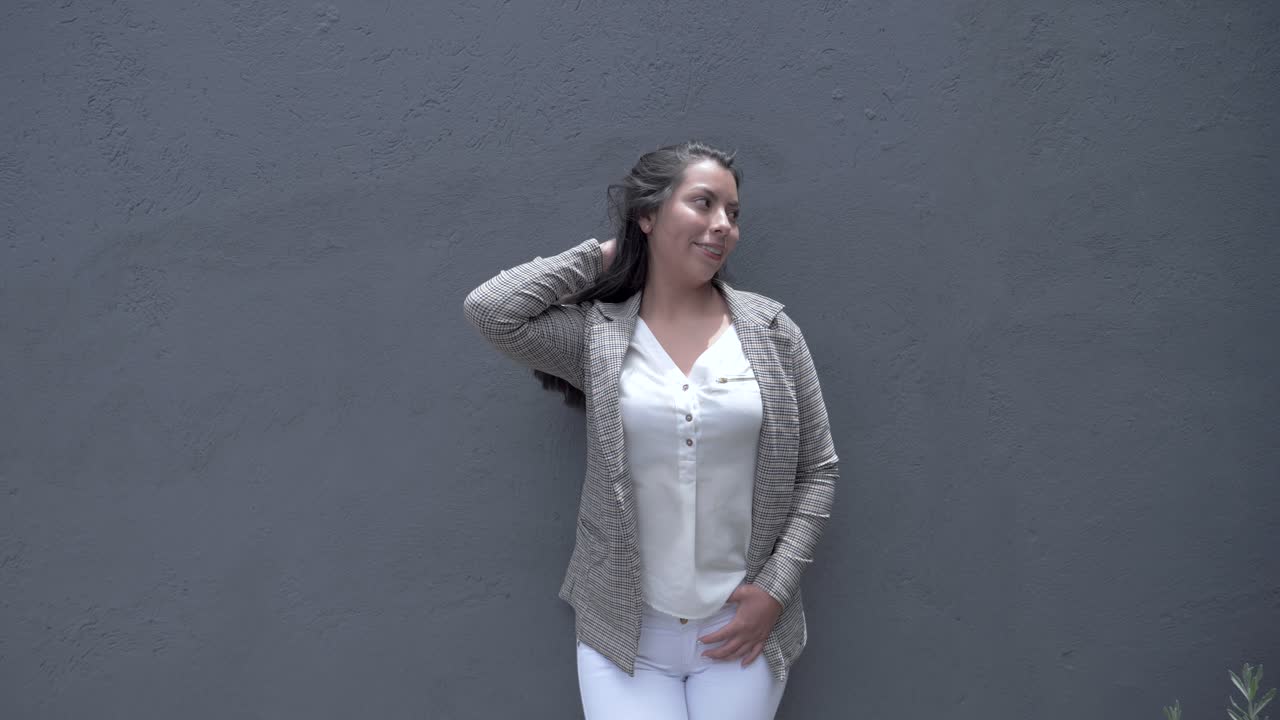 chica feliz apoyada en una pared gris, sonriendo mientras se arregla el cabello, mostrando confianza y felicidad en su apariencia y comportamiento